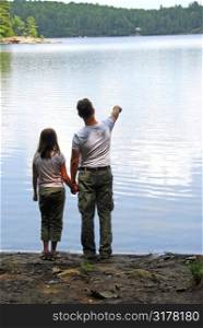 Father and daughter standing on the lake shore and looking at calm water, father pointing at somthing on the other shore