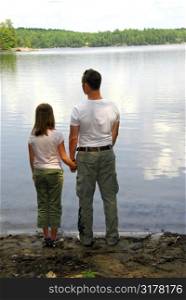 Father and daughter standing on the lake shore and looking at calm water