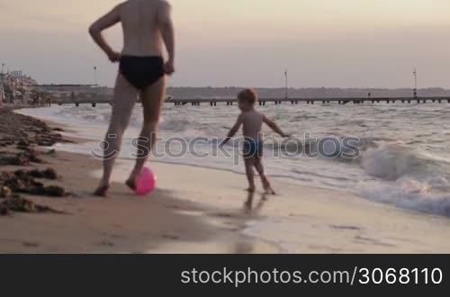 Father and his little son running along the beach playing ball in their swimsuits in evening light moving away from the camera along the sand at the edge of the surf