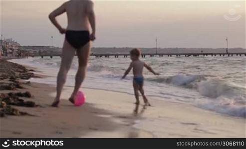 Father and his little son running along the beach playing ball in their swimsuits in evening light moving away from the camera along the sand at the edge of the surf
