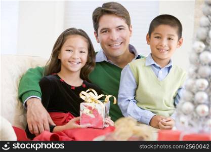 Father And His Son And Daughter Sitting On Sofa,Holding A Christmas Present