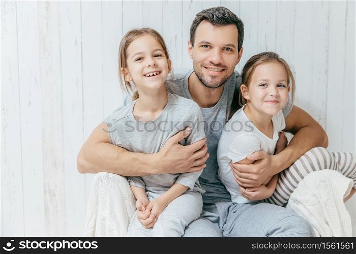 Father and kids. Paternity concept. Handsome unshaven affectionate dad embraces his two pretty dauhters, going to have walk together, have good relationship, pose at camera with positive smiles