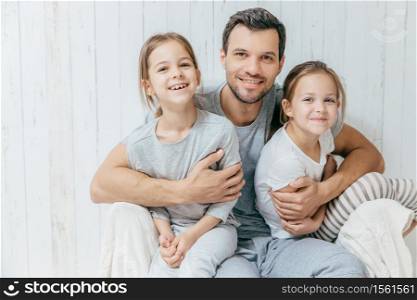 Father and kids. Paternity concept. Handsome unshaven affectionate dad embraces his two pretty dauhters, going to have walk together, have good relationship, pose at camera with positive smiles