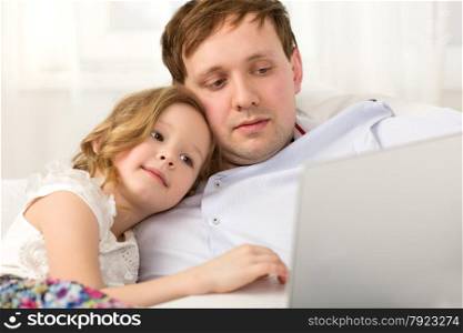 Father and little daughter on the sofa at home. Father holding laptop, girl using it leaning on dads shoulder