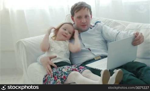 Father and lovely daughter lying on the sofa with laptop. Dad telling girl something. Leisure time with family