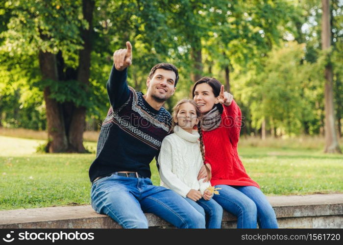 Father and mother sit between their daughter against trees or nature background, show something to her, indicate with fore fingers. Smiling parents show bird to little beautiful girl. Parenthood