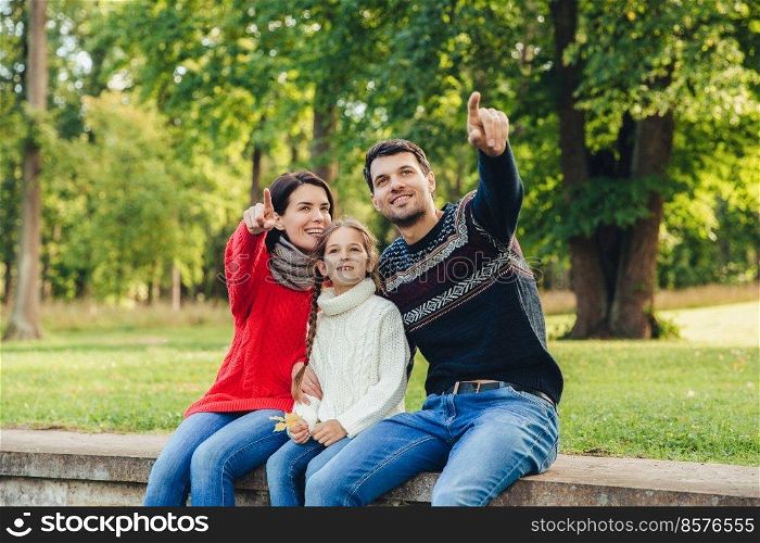 Father and mother sit between their daughter against trees or nature background, show something to her, indicate with fore fingers. Smiling parents show bird to little beautiful girl. Parenthood