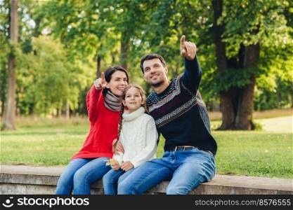 Father and mother sit between their daughter against trees or nature background, show something to her, indicate with fore fingers. Smiling parents show bird to little beautiful girl. Parenthood