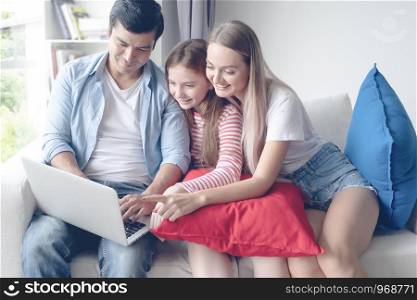 father and mother with daughter smiling and sitting on sofa together and using internet laptop at home, happy young family concept