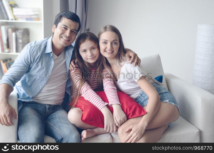 father and mother with daughter smiling and sitting on sofa together at home, happy young family concept