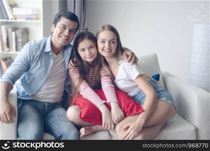 father and mother with daughter smiling and sitting on sofa together at home, happy young family concept