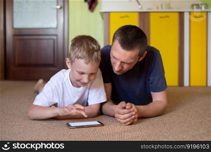 father and son, boy with dad, look at the phone, speakerphone, call mom, watch cartoons, play phone, gadget, lie on the floor, games at home, time with dad. A cute boy lies with his dad and looks at the phone