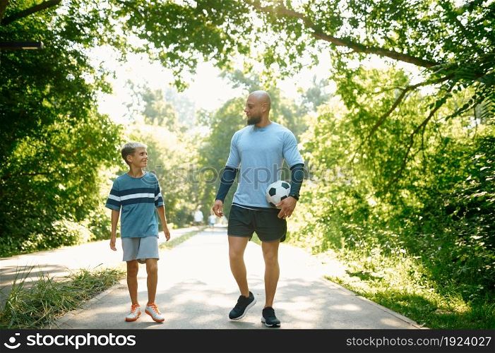 Father and son, football players on walking path outdoors. The family leads a healthy lifestyle, daddy and boy with ball in summer park. Father and son, football players on walking path