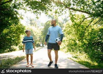 Father and son, football players on walking path outdoors. The family leads a healthy lifestyle, daddy and boy with ball in summer park. Father and son, football players on walking path