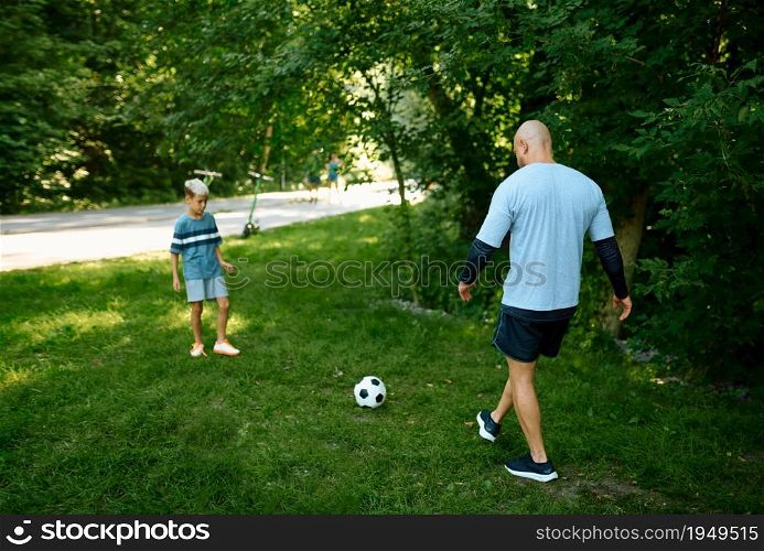 Father and son play football on the grass outdoors. The family leads a healthy lifestyle, morning fitness workout in summer park. Father and son play football on the grass outdoors