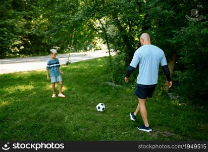 Father and son play football on the grass outdoors. The family leads a healthy lifestyle, morning fitness workout in summer park. Father and son play football on the grass outdoors
