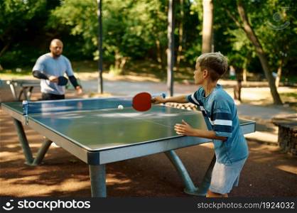 Father and son play table tennis outdoors. The family leads a healthy lifestyle, daddy and boy, ping pong training in summer park. Father and son play table tennis outdoors