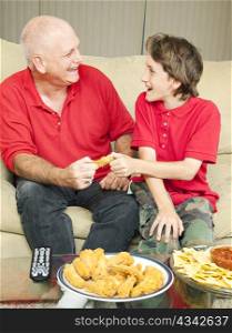 Father and son playfully fighting over a chicken wing as they watch the football game.