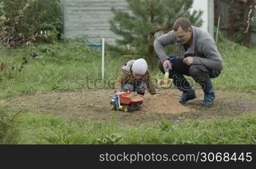 Father and son playing with toy truck loading it with sand. Family fun outdoors.