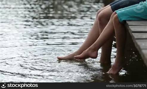 Father and son sitting on wooden pier dangling their legs into the water and splashing the lake on warm summer day. Side view. Happy dad and teenage boy having fun on pier splashing their legs into pond water.