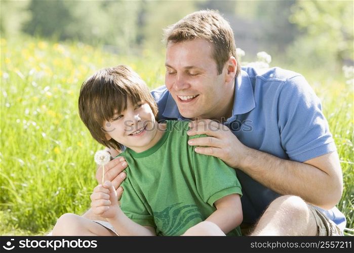 Father and son sitting outdoors with dandelion head smiling