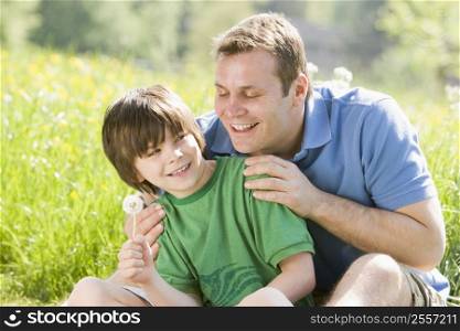 Father and son sitting outdoors with dandelion head smiling