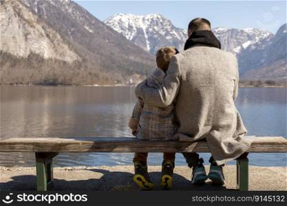 Father and son sitting together outdoors on the bench and enjoying mountains, snow, good weather, blue sky. Little boy and his father spending time together. Family time. Beautiful winter landscape. Father and son sitting together outdoors on the bench and enjoying mountains, snow, good weather, blue sky. Little boy and his father spending time together. Family time. Beautiful winter landscape.