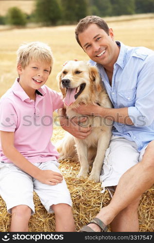 Father And Son Sitting With Dog On Straw Bales In Harvested Field