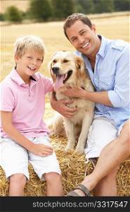 Father And Son Sitting With Dog On Straw Bales In Harvested Field