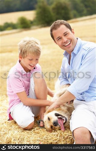 Father And Son Sitting With Dog On Straw Bales In Harvested Field