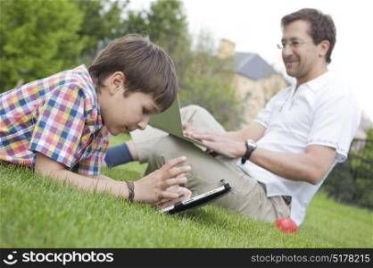 Father and son using laptop and tablet lying on the grass