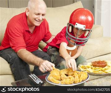 Father and son watching football together and eating snacks.