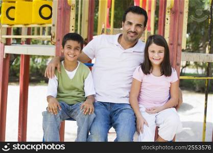 Father and two children sitting on playground structure smiling (selective focus)