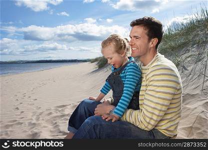 Father embracing daughter (5-6) sitting on beach