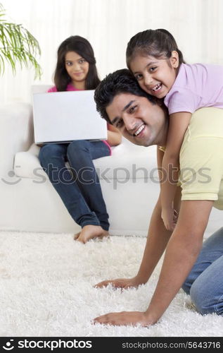 Father giving daughter a ride on his back with woman using laptop in the background