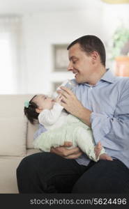 Father holding and feeding his baby daughter on the couch