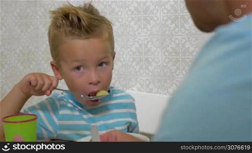 Father is teaching blond son how to use a spoon during eating some fruit mix from plate