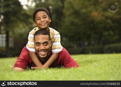 Father lying in grass smiling as son climbs on his back and hugs his neck.