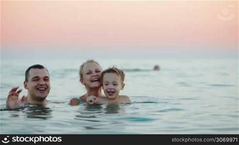 Father, mother and little son bathing in sea at sunset and waving with their hands