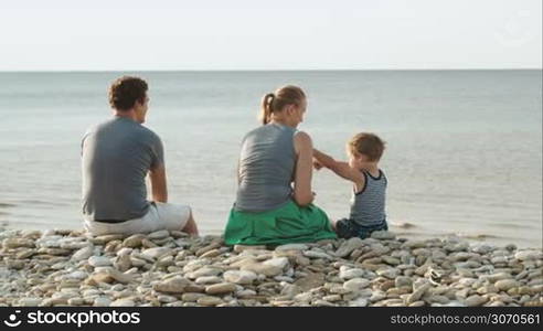 Father, mother and son sitting on pebble beach by the sea. Woman and child playing, man enjoying the view, boy showing something in water