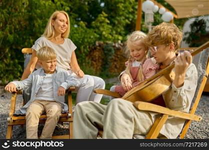 Father plays guitar for his family at the trailer, summer camping. Family with children travel in camp car, nature and forest on background. Campsite adventure, travelling lifestyle, motorhome. Father plays guitar for his family at the trailer