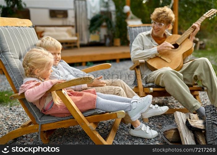 Father plays guitar for kids at the trailer, summer camping. Family with children travel in camp car, nature and forest on background. Campsite adventure, travelling lifestyle. Father plays guitar for kids at trailer, camping