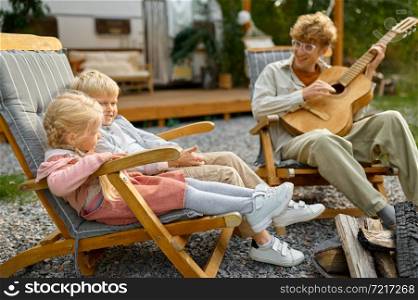 Father plays guitar for kids at the trailer, summer camping. Family with children travel in camp car, nature and forest on background. Campsite adventure, travelling lifestyle. Father plays guitar for kids at trailer, camping