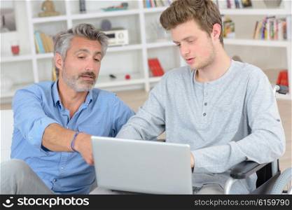 Father pointing to screen of laptop held by son in wheelchair