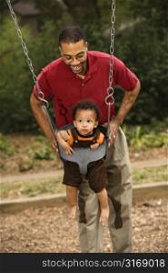 Father pushing young son on swing in park in playground.