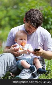 Father sitting on green grass using a mobile phone reading a text message with a baby with a dummy in its mouth on his lap
