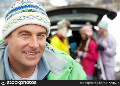 Father Smiling At Camera Whilst Family Load Skis In Boot Of Car