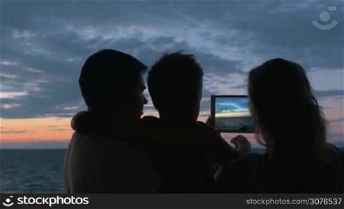 Father stands with son in his arms, and mother is photographed on tablet of sunset and sea in city Perea, Greece. Then mom laughed and kisses his son
