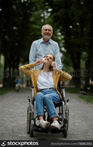 Father walks with disabled daughter in wheelchair, family in summer park. Paralyzed people and disability, handicap overcoming. Handicapped female person and adult male guardian in public place. Father walks with disabled daughter in wheelchair