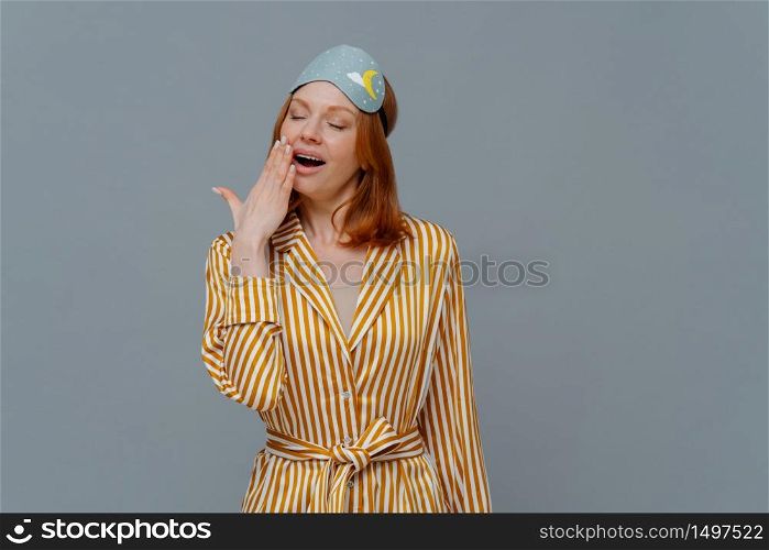 Fatigue sleepy red head woman yawns with opened mouth, wears domestic striped robe and blindfold on head, wants to sleep, stands against grey background, feels weak. Late night time concept.